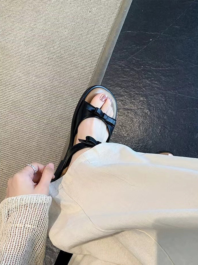 A person wearing a black flat sandal with a wide strap and bow detail, paired with light beige wide-leg pants. The photo is taken from a top-down perspective, showing a hand adjusting the pant leg and the textured floor beneath.