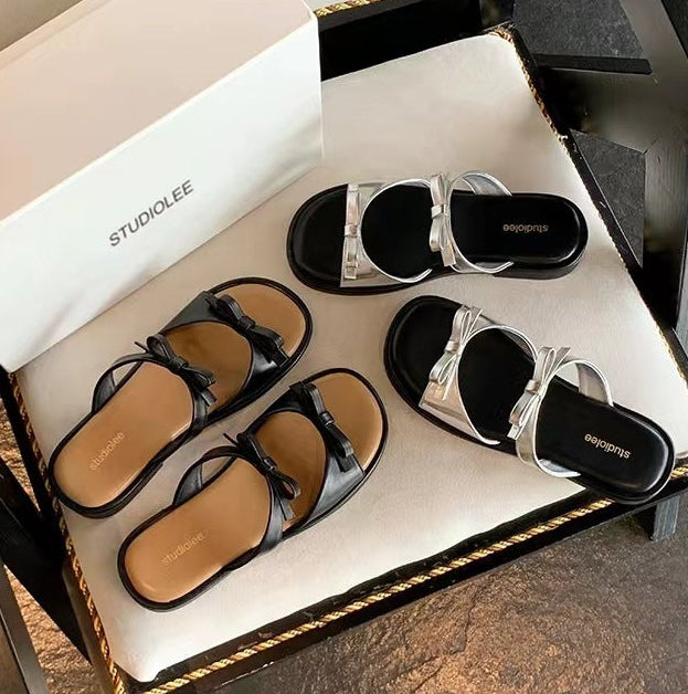 A pair of black slide sandals with metallic silver straps, displayed on a beige cushioned surface next to their white box. The image includes Chinese promotional text above and below the product photo.