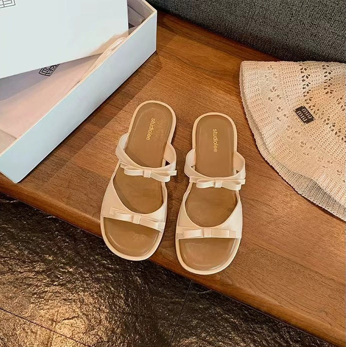 A pair of cream-colored slide sandals with bow details, placed on a wooden surface next to a shoe box and a woven textile.