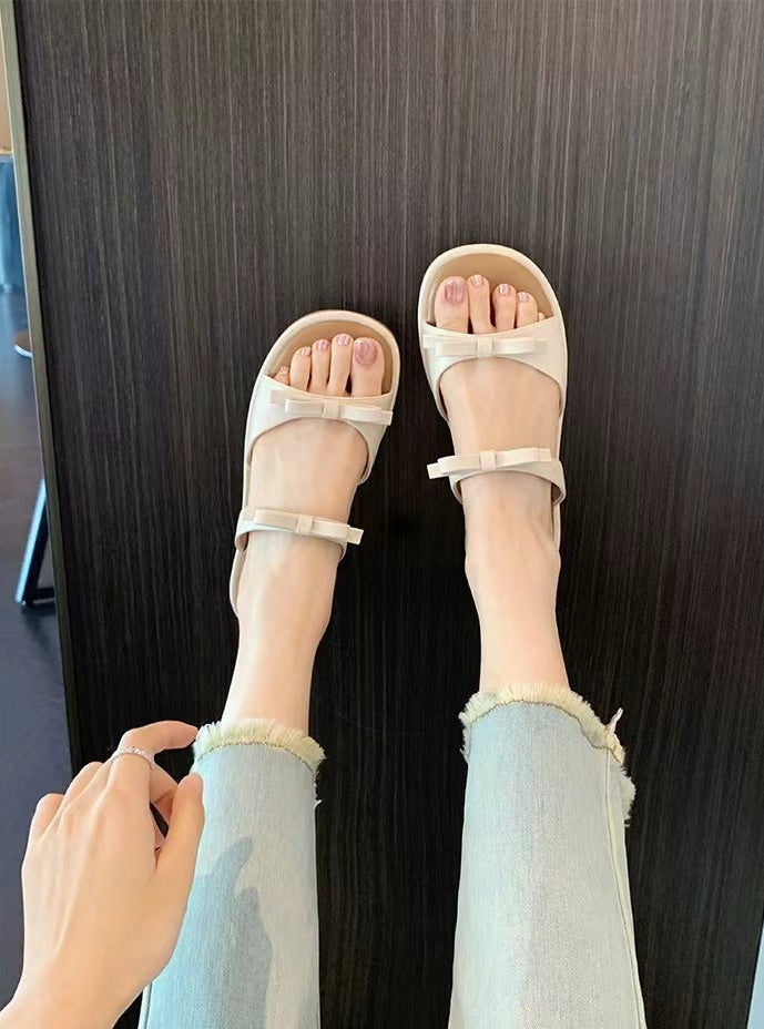 A person wearing light beige slide sandals with bow details and frayed-jeans legs, photographed from above against a dark wood floor.