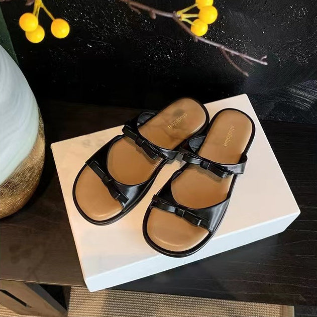 A pair of black leather slide sandals with brown insoles, displayed on a white rectangular box against a dark wooden background with decorative yellow berries and a ceramic vase.