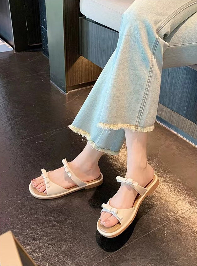 A person wearing light beige slide sandals with bow details and frayed denim jeans, standing on a dark tiled floor in an indoor setting. Chinese text is visible at the bottom of the image.