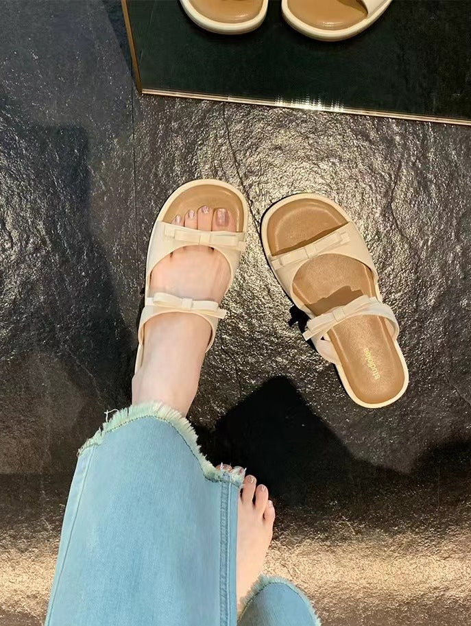 A person wearing beige double-strap slide sandals with frayed light-wash jeans, standing on a dark textured floor. A mirror at the top shows the sandals from above.