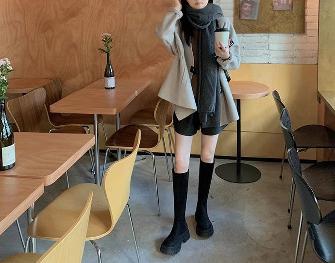 A woman stands in a cafe wearing a light beige trench coat, a dark gray scarf, black shorts, and black knee-high socks.