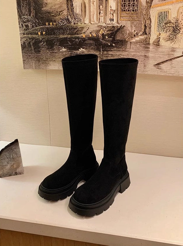 A pair of black knee-high boots with chunky lug soles, displayed on a white shelf against a beige wall with a framed print in the background.