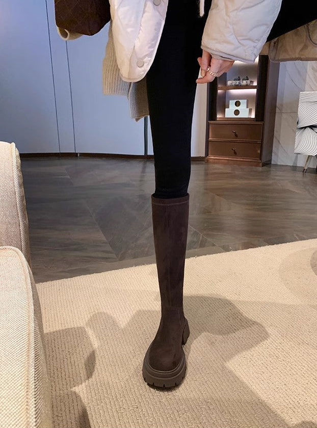 A person wearing a long dark brown knee-high boot with a chunky sole, paired with black leggings and a white puffer jacket, standing in a modern indoor room with a visible text overlay at the bottom.