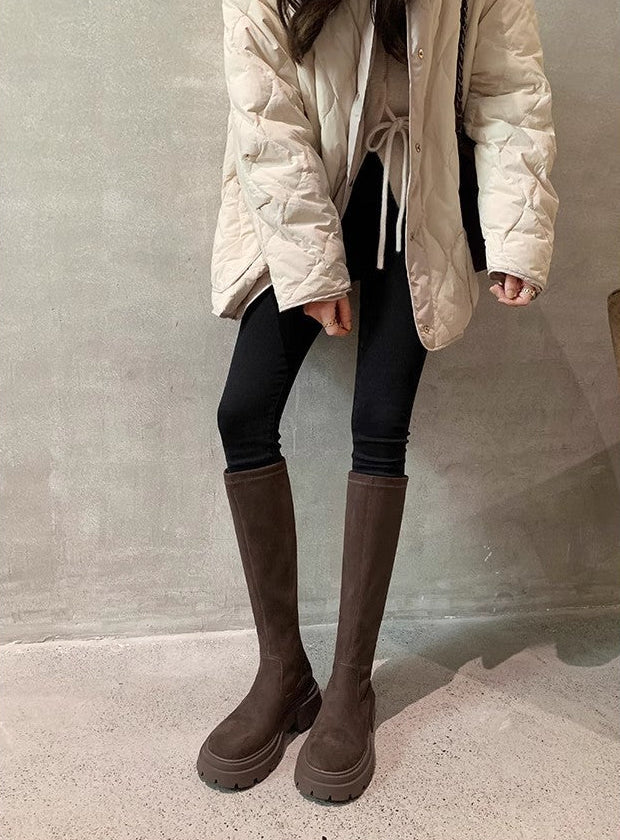 A model wearing a light beige quilted puffer jacket, black leggings, and dark brown knee-high boots with thick platform soles, standing against a textured gray wall.