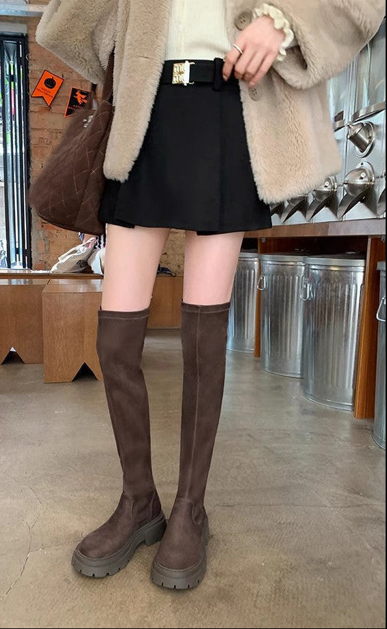 A person standing indoors wearing knee-high brown suede-like boots with chunky soles, a black mini skirt, a cream top, and a beige fuzzy coat, with a brown shoulder bag in a shop interior.