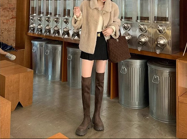 A woman stands in a retail store wearing a beige coat, black skirt, and brown knee-high boots, holding a brown handbag. The background features a wooden wall with framed landscape prints and stainless steel dispensers.