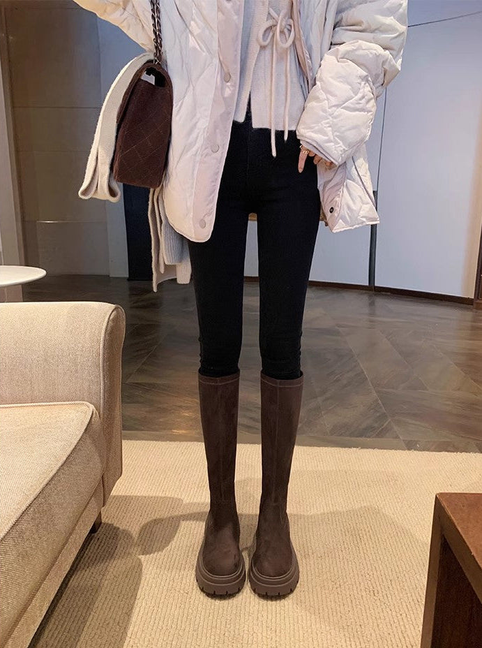 A person standing indoors wearing knee-high dark brown boots, black skinny pants, a white puffer jacket, and carrying a dark brown shoulder bag.