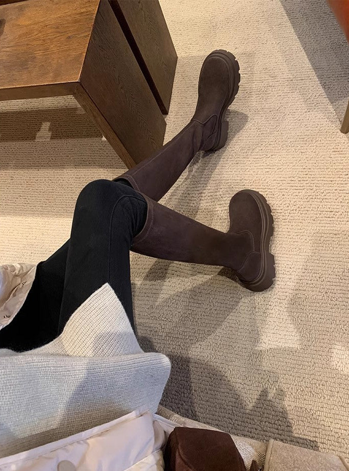 A person wearing dark brown knee-high socks or over-the-knee socks with matching dark brown ankle boots, seated on a sofa in a home setting with a wooden table and carpet visible.