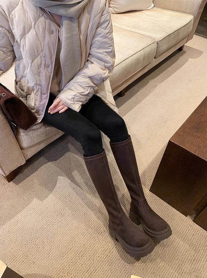 A person seated on a beige sofa wearing dark brown knee-high boots, black leggings, and a light beige puffer jacket in a living room setting.