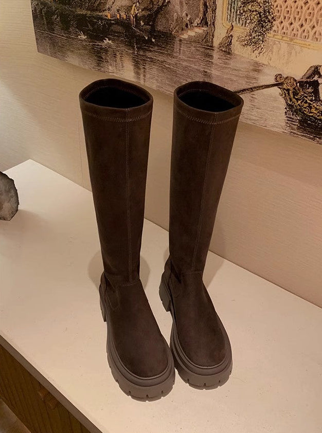A pair of dark brown mid-calf boots with chunky lug soles, displayed on a white shelf against a beige wall with a framed print.