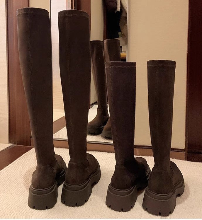 Four dark brown over-the-knee boots with chunky platform soles, shown from the back in a row on a light carpet in front of a mirror.