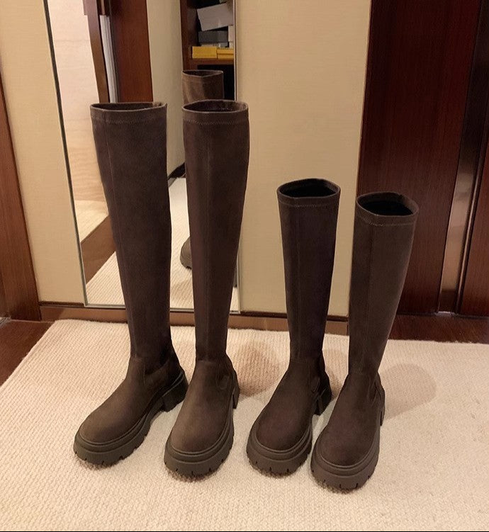 Four dark brown knee-high boots in different heel heights arranged on a light beige rug in front of a mirror and wooden door.